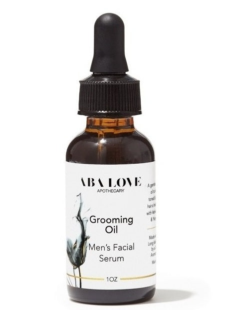 Amber glass bottle with black dropper. It has a white label with black and grey graphic and black type. Grooming Oil Men’s Facial Serum, 1 oz bottle. There is a thin gold label printed around the bottom of the label.