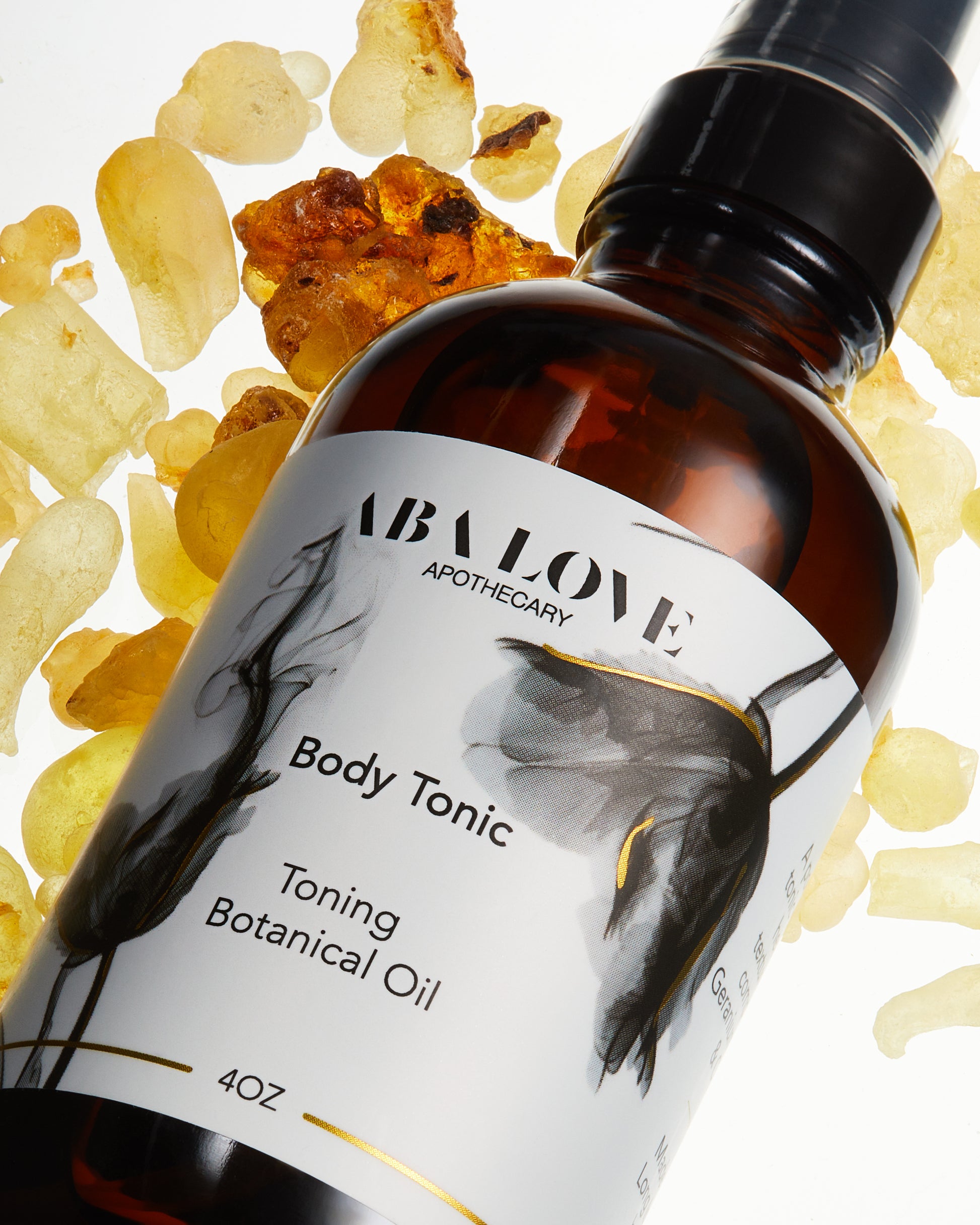 Amber colored bottle with black treatment pump.  "Aba Love Apothecary Body Tonic Toning Botanical Oil". The label is white with gold trim and grey flowers. The bottle lays flat on pieces of yellow colored frankincense resin