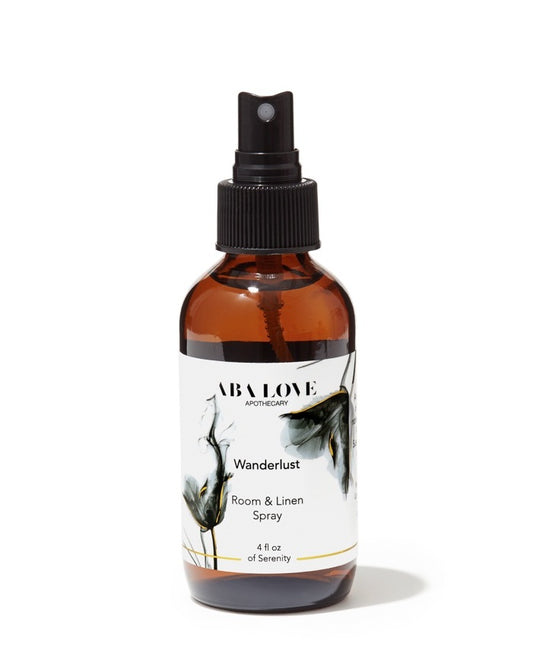 Amber glass bottle with black sprayer mist top. The amber glass bottle has a white label with black and grey graphic and black type that reads Wanderlust Room and Linen Spray,  4 oz bottle.There is a thin gold line printed around the bottom of the label.